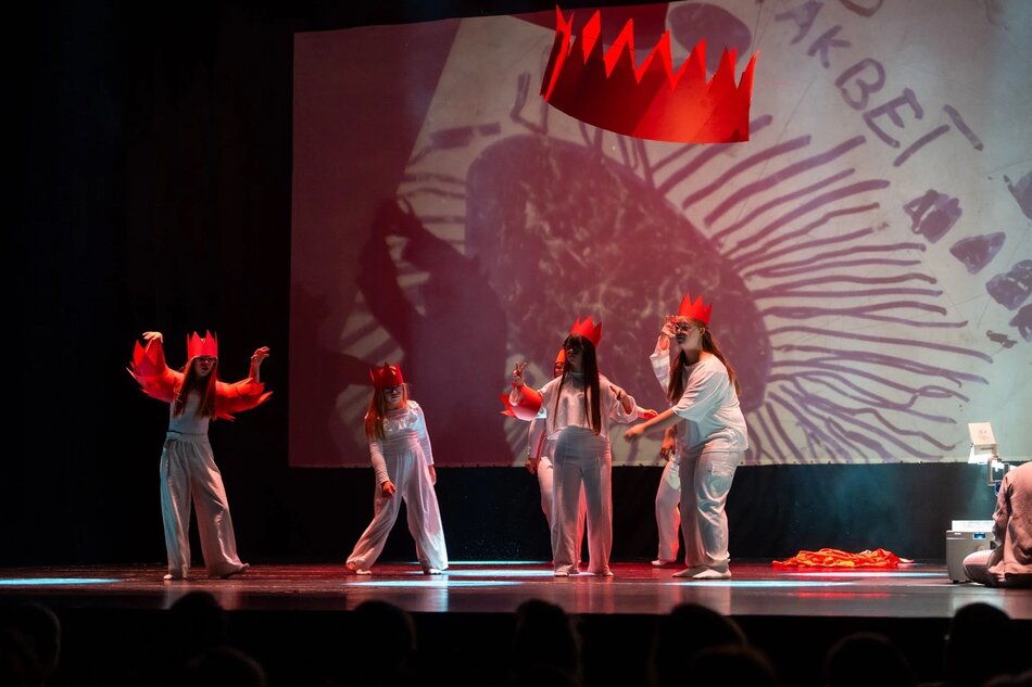 Zdjęcie przedstawia scenę teatralną, na której młodzi aktorzy w białych strojach i czerwonych koronach odgrywają dynamiczną scenę na tle dużej projekcji z napisem „MAKBET” i rysunkiem przypominającym oko. Nad nimi unosi się zawieszona czerwona korona, a całość ma ekspresyjny i symboliczny charakter, nawiązujący do dramatu Szekspira