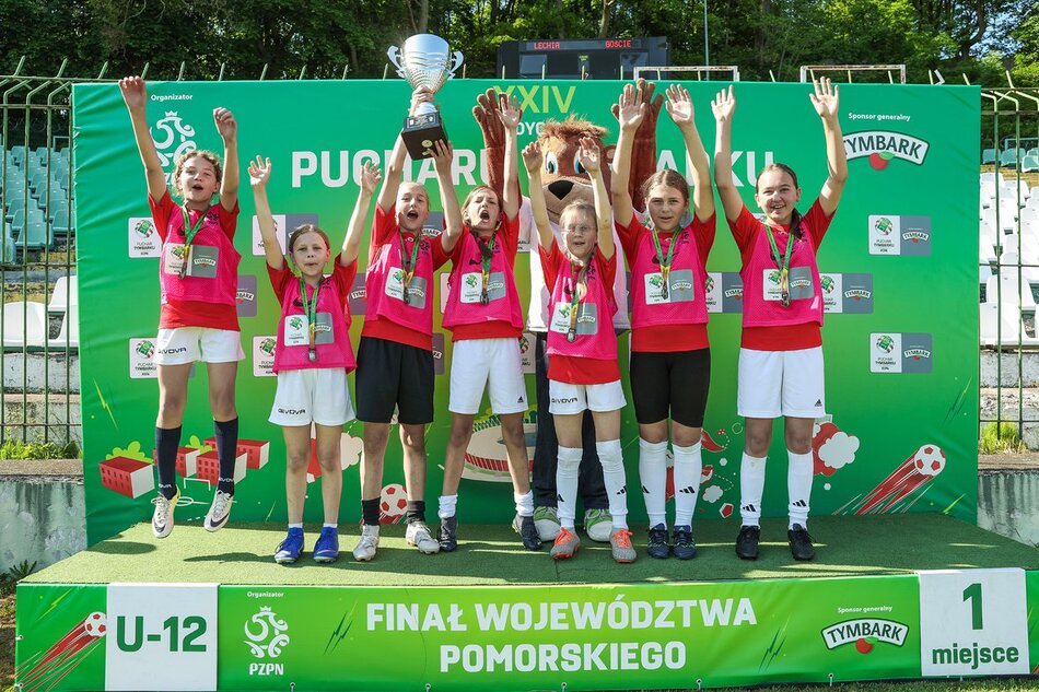 Drużyna dziewcząt U-12 świętuje zdobycie 1. miejsca w finale województwa pomorskiego Pucharu Tymbarku, trzymając puchar i medale na podium. 