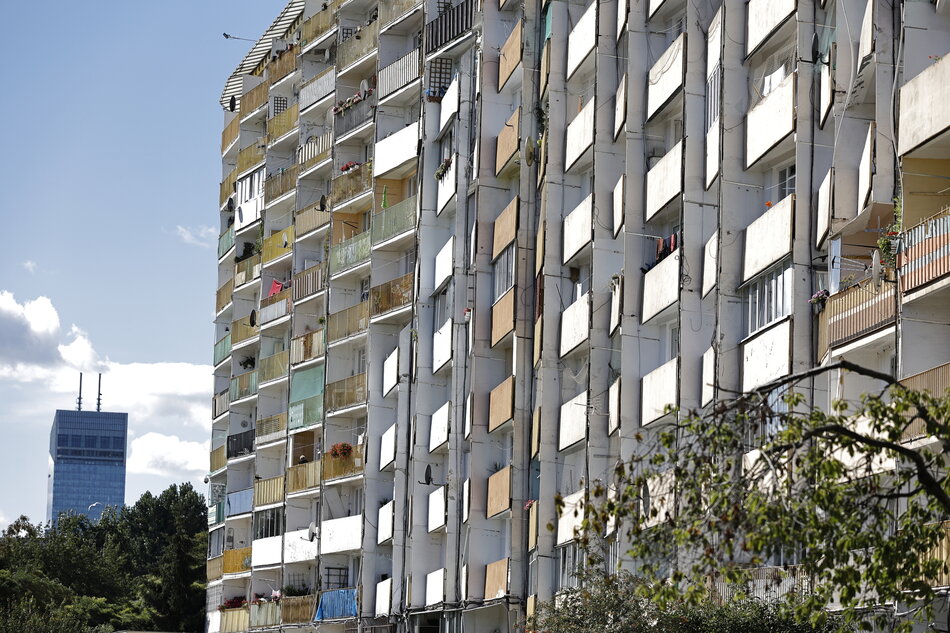 Zdjęcie przedstawia typowe osiedle mieszkaniowe z wielkiej płyty – prawdopodobnie w dzielnicy Przymorze w Gdańsku. Widać charakterystyczny dla tego obszaru długi blok mieszkalny, z licznymi balkonami i zróżnicowanymi kolorystycznie osłonami przeciwsłonecznymi. Balkony są zróżnicowanie zagospodarowane – część z nich jest zadaszona, część oszklona lub ozdobiona kwiatami. W tle, po lewej stronie, widoczny jest nowoczesny, wysoki budynek z dwoma masztami na dachu – może to być Olivia Star lub inny biurowiec z kompleksu Olivia Business Centre.