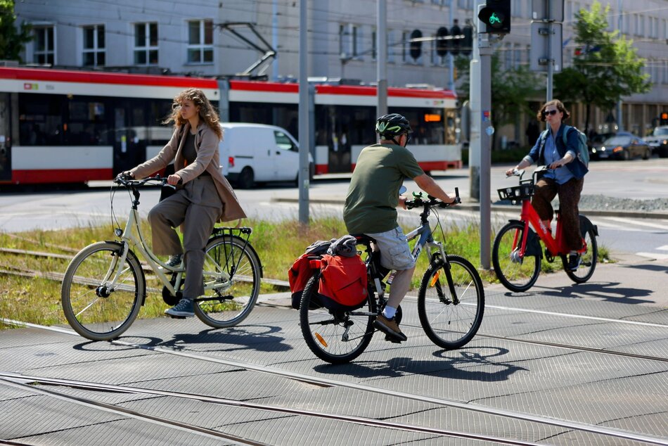 Na zdjęciu widać grupę osób jadących na rowerach po wyznaczonej ścieżce rowerowej w miejskim otoczeniu. Pogoda jest słoneczna, a rowerzyści są ubrani lekko,