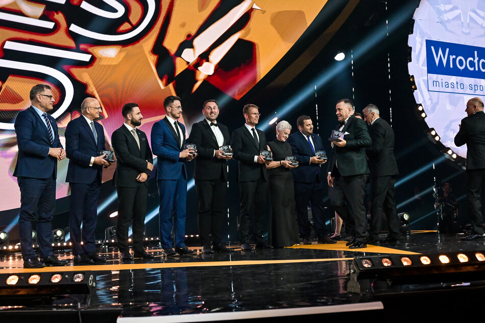 Grupa elegancko ubranych osób odbiera nagrody na uroczystej gali, odbywającej się na efektownie oświetlonej scenie. 
