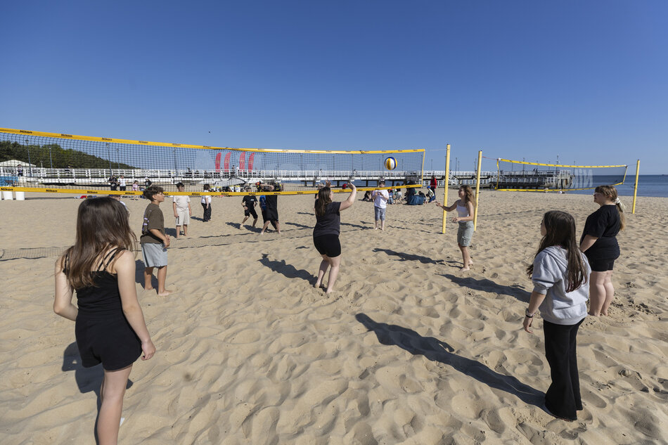  grupa młodych ludzi grających w siatkówkę na piaszczystej plaży. Jest kilku graczy na pierwszym planie, niektórzy stoją, a niektóre chodzą po dziedzińcu. dziedziniec jest urządzony z żółtą siatkę i między dwoma graczami jest czerwono-biała piłka siatkowa. w tle jest kilka osób siedząc na ławkach i oglądając mecz. po lewej stronie stoi z tyłu do kamery, patrząc w górę na piłkę.
