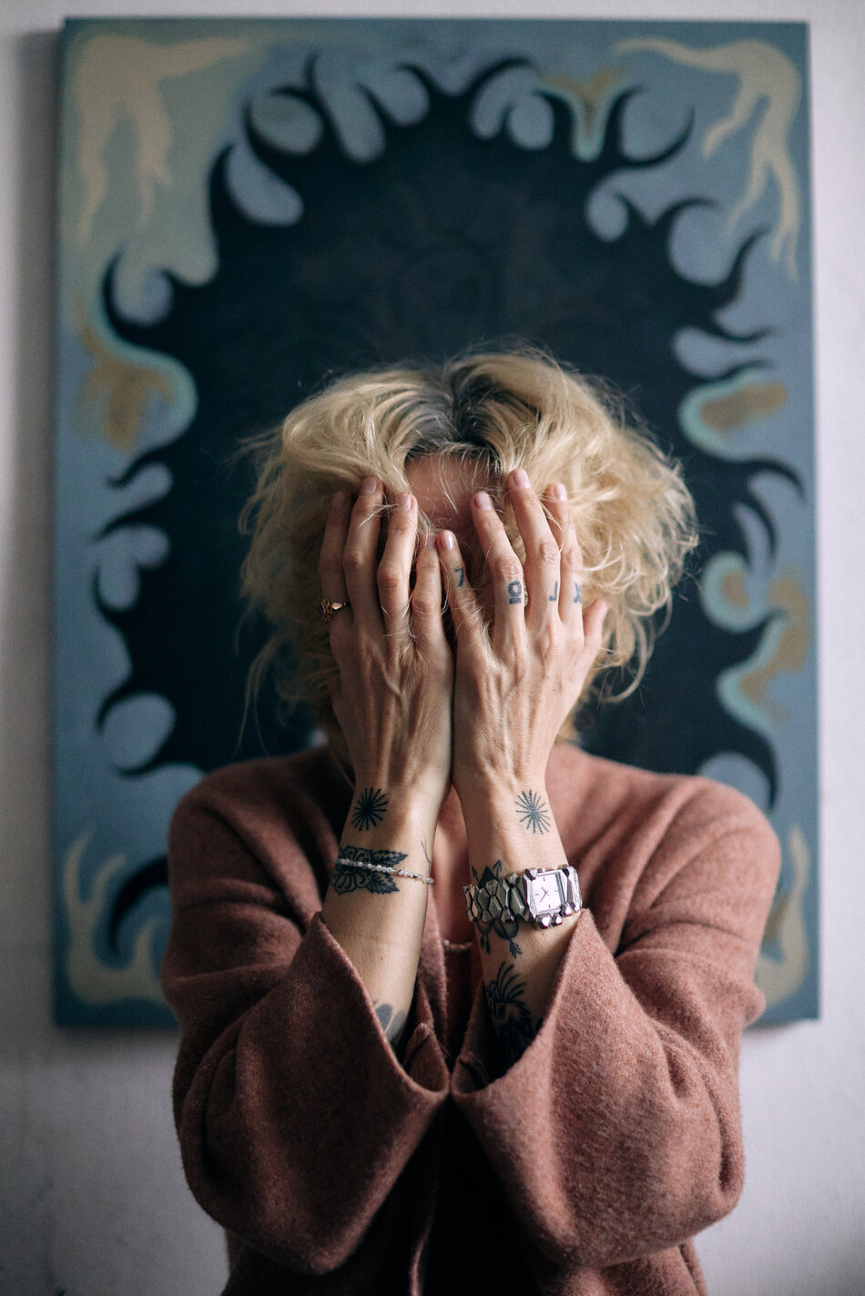 Na zdjęciu kobieta zakrywająca twarz rękoma, w tle obraz z czarnymi elementami.
