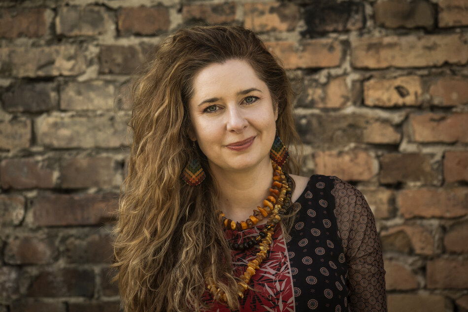 Na zdjęciu uśmiechnięta kobieta z rozpuszczonymi za ramiona włosami, z naszyjnikiem z bursztynów. W tle ściana z cegieł.