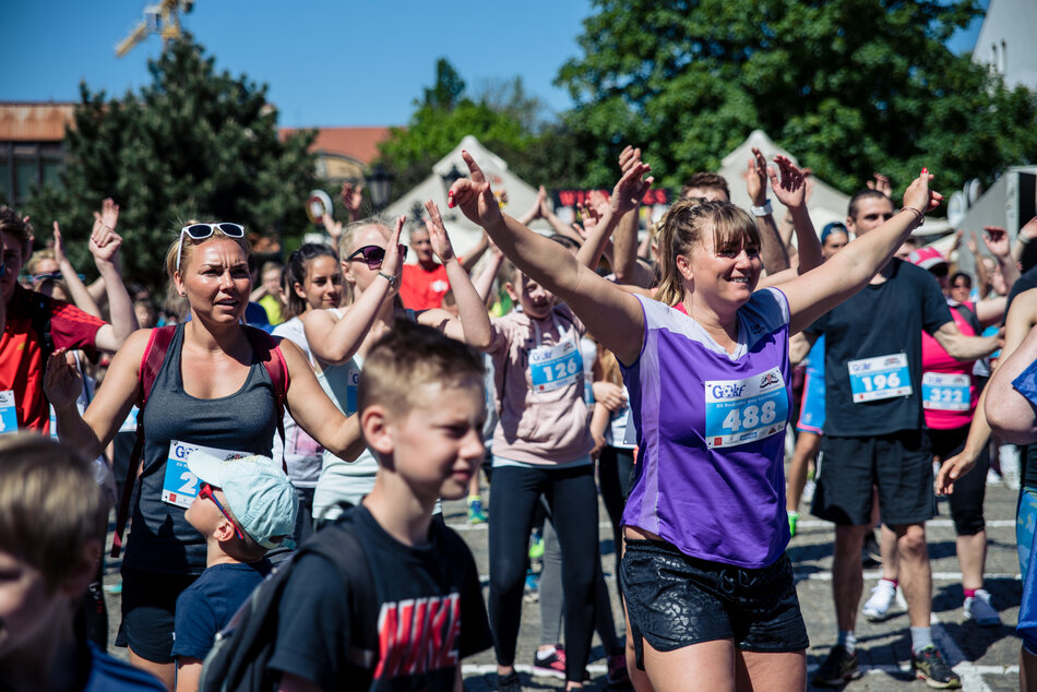 Na zdjęciu widać grupę uczestników rozgrzewających się przed biegiem, unoszących ręce w rytm ćwiczeń rozgrzewkowych, w słoneczny dzień. Wszyscy mają przypięte numery startowe, a atmosfera wydaje się radosna i pełna energii.