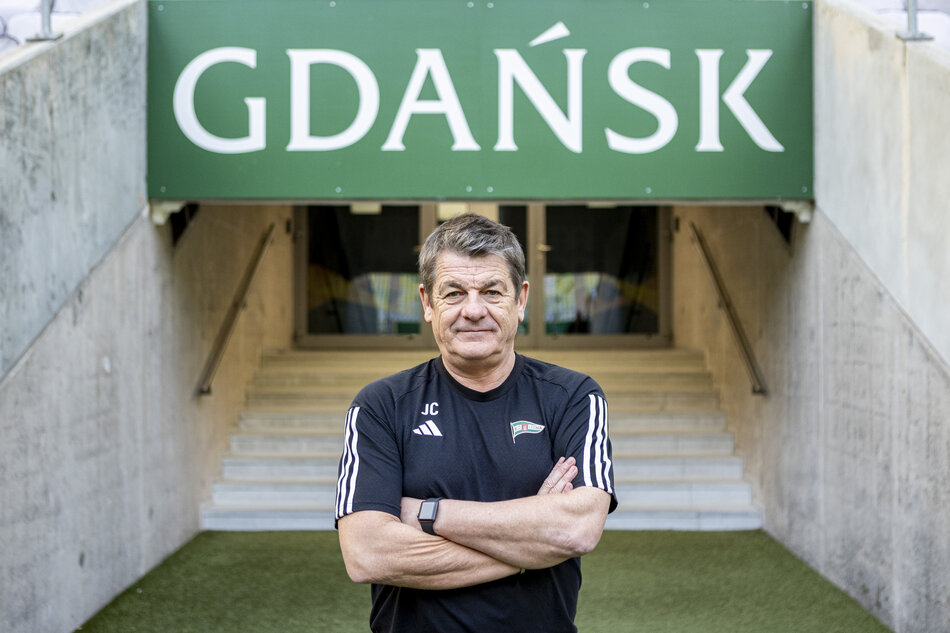 Trener w sportowym stroju stoi z założonymi rękami przed wejściem na stadion w Gdańsku. 
