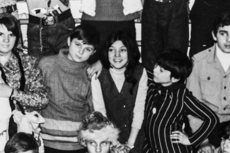 Na czarno-białym zdjęciu widzimy grupę dzieci, prawdopodobnie uczniów szkoły podstawowej, pozujących do wspólnej fotografii klasowej lub pamiątkowej. W centralnej części zdjęcia znajdują się trzy dzieci: chłopiec w swetrze z golfem, dziewczynka w kamizelce w paski oraz dziewczynka w pasiastym stroju z ręką opartą na biodrze. W tle widać inne dzieci, niektóre częściowo zasłonięte.