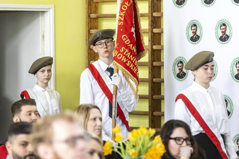 Na zdjęciu widzimy uroczystość szkolną, najprawdopodobniej akademię lub święto patriotyczne. Centralną postacią jest uczeń trzymający sztandar z nazwą szkoły: Szkoła Podstawowa nr 69 w Gdańsku im. gen. Stanisława Sosabowskiego. Obok niego stoją dwie inne osoby, również w strojach galowych i z szarfami, co sugeruje, że pełnią funkcję pocztu sztandarowego.