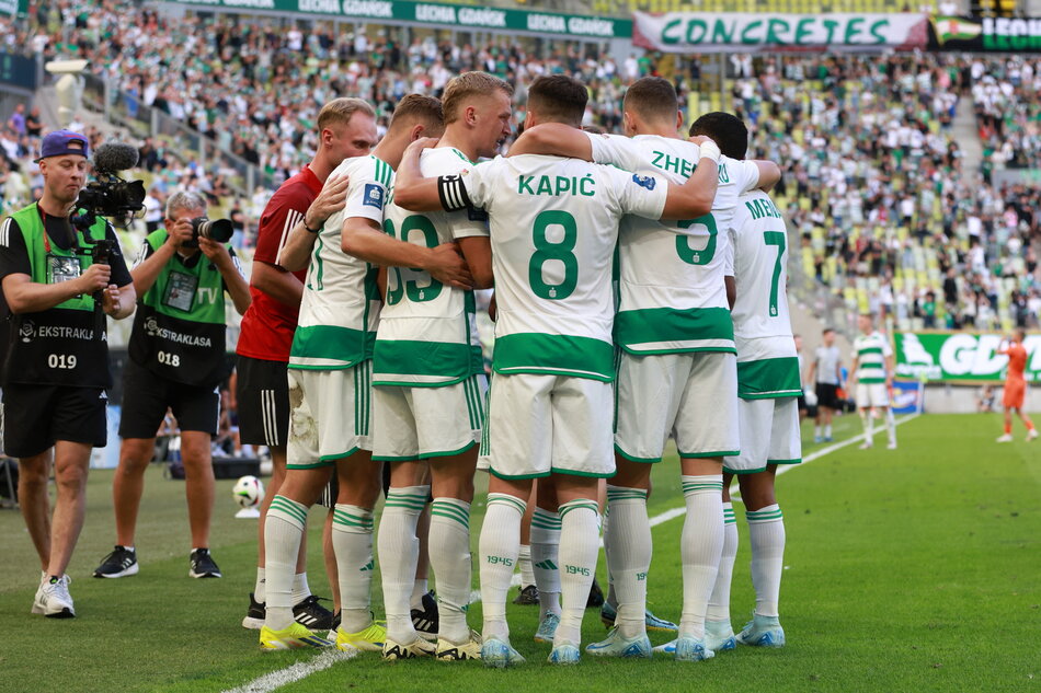  grupa piłkarzy zebranych razem w kręgu na boisku, noszą zielone i białe koszulki z numerami 8 i 7 na nich, gracze stoją blisko siebie, z ramionami wokół swych ramion, w tle jest duży tłum widzów na trybunach, po lewej stronie obrazu można zobaczyć kamerzystę trzymającego kamerę wideo i rejestrującego moment.