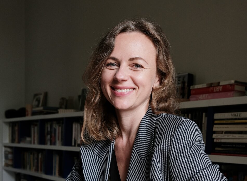 Na zdjęciu kobieta w lekko kręconych włosach, w szarej marynarce. W tle regał z książkami.