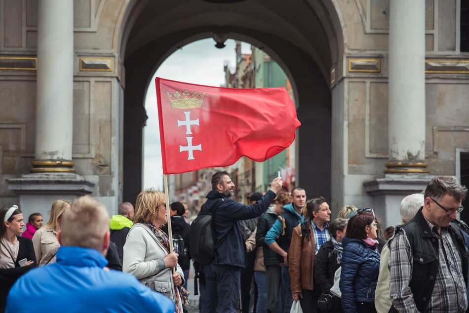 Kobieta z flagą Gdańska stoi przed Złotą Bramą, w tłumie przechodzących ludzi