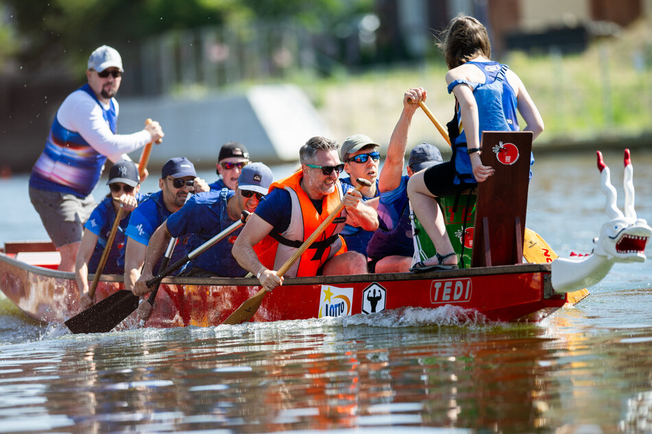 Zespół intensywnie wiosłuje w wyścigu smoczych łodzi, skupiony i zdeterminowany, płynąc po spokojnej wodzie. 