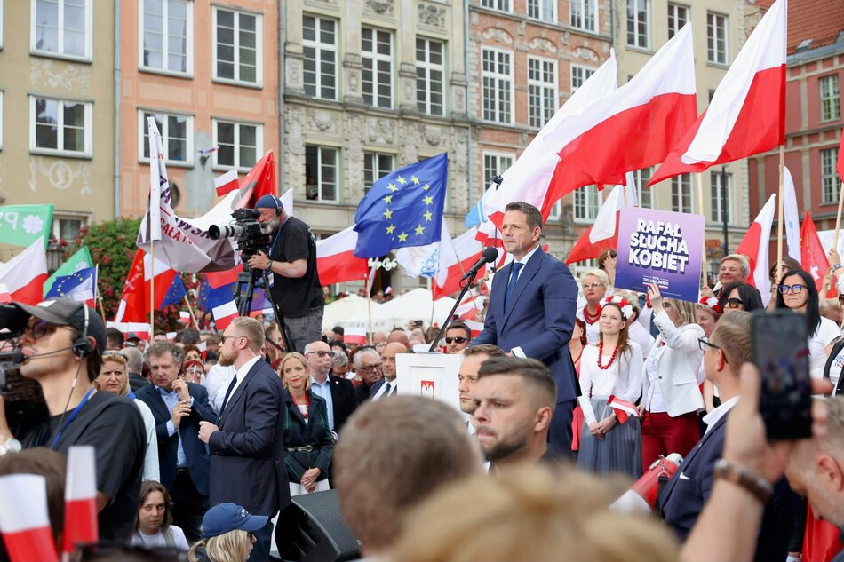 Na zdjęciu widzimy wiec polityczny w Polsce. W centrum znajduje się mężczyzna przemawiający z mównicy — prawdopodobnie kandydat lub polityk — otoczony przez tłum ludzi, wielu z nich trzyma polskie flagi. W tle widać również flagi Unii Europejskiej oraz baner z napisem „Rafał słucha kobiet” (co można odczytać jako hasło wspierające polityka o imieniu Rafał, być może Rafała Trzaskowskiego).
