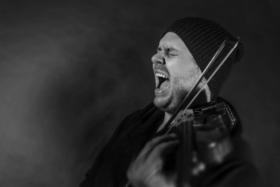 Na zdjęciu mężczyzna z szeroko otwartymi ustami, czapką na głowie, grający na skrzypcach.