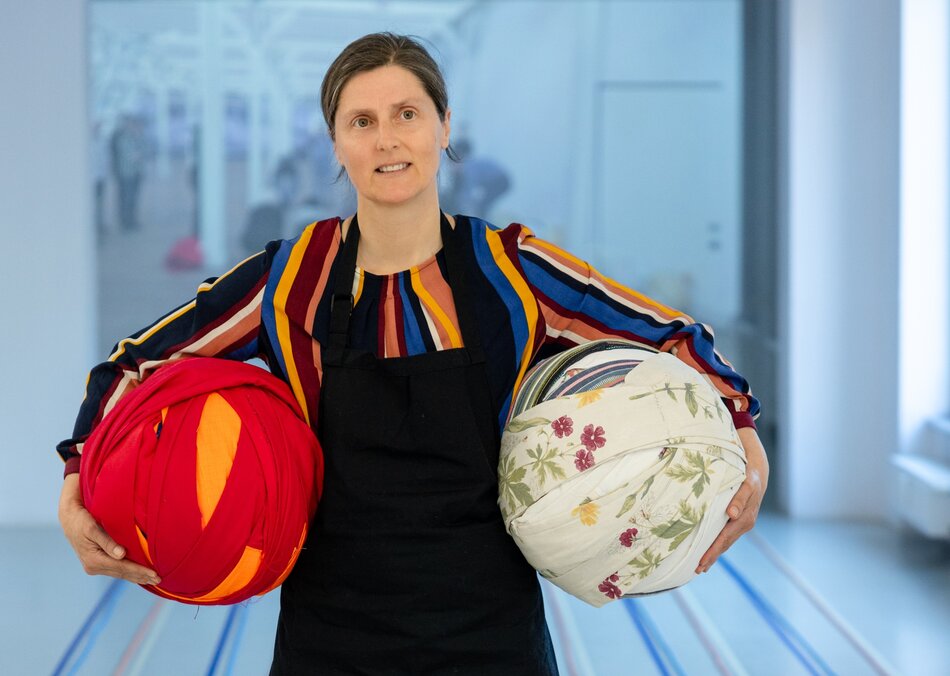 Na zdjęciu kobieta w czarnej sukience, z kolorowymi paskami. Trzyma w rękach dwie duże kolorowe kule.