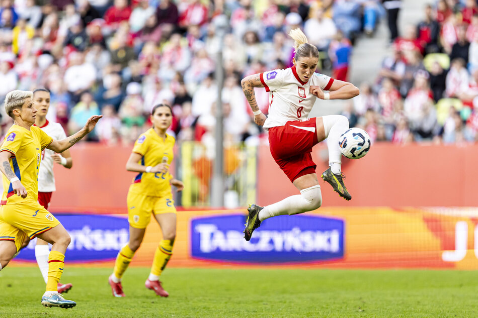 młoda kobieta w dwuczęściowym stroju sportowym wyskakuje do piłki, w trakcie meczu na dużym stadionie. obok widoczne zawodniczki drużyny przeciwnej, w tle kibice na trybunach