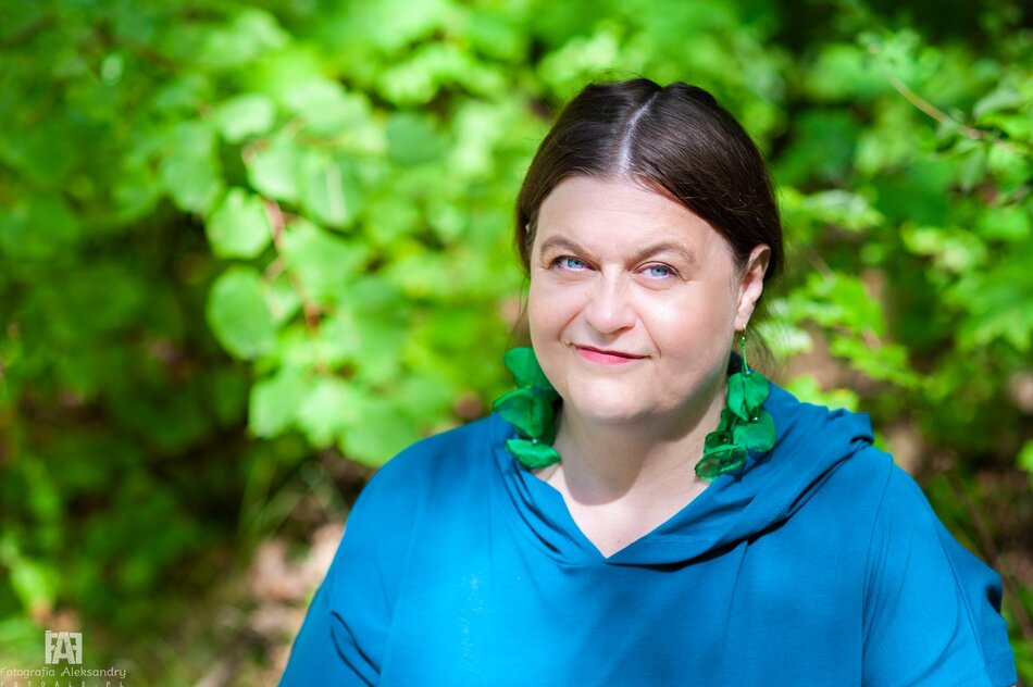 Na zdjęciu kobieta ubrana w niebieską sukienkę, z zielonymi kolczykami, na tle zielonych soczyście liści.