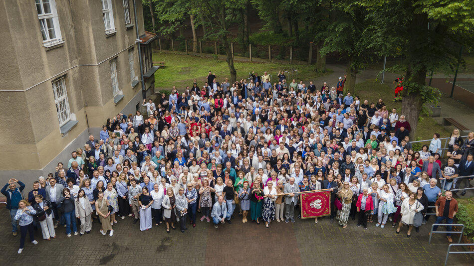 Duża grupa osób stoi na dziedzińcu szkoły i pozuje do zbiorowej fotografii robionej z lotu ptaka, z drona.