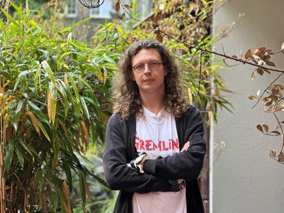 Na zdjęciu mężczyzna w okularach, z długimi włosami za ramiona, w tle zielona roślinność.