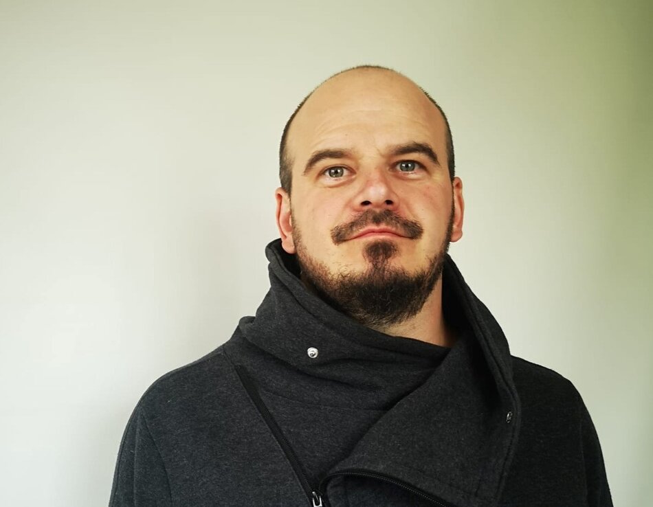 Na zdjęciu mężczyzna w czarnej bluzie, na twarzy widoczny zarost.