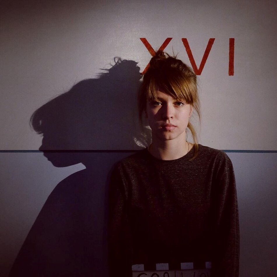 Na zdjęciu kobieta z upiętymi włosami, w tle rzymskie cyfry 16 oraz cień kobiety.