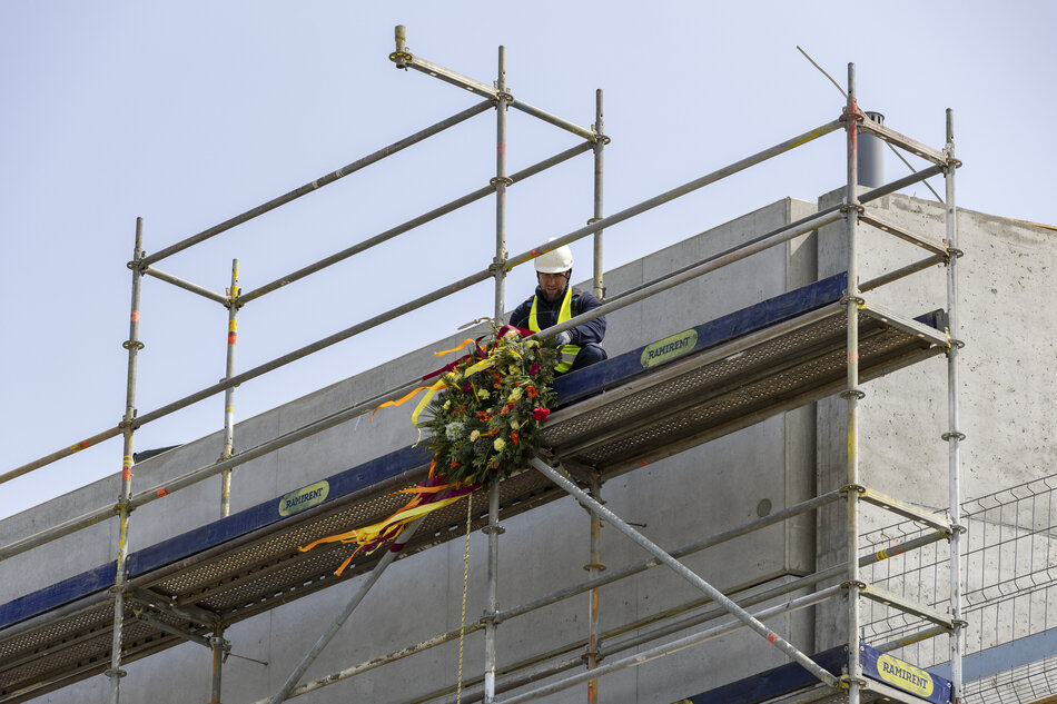 Na zdjęciu widzimy mężczyznę w kasku ochronnym i kamizelce odblaskowej, który umieszcza wiechę — tradycyjny wieniec z gałęzi i kwiatów — na rusztowaniu budynku w trakcie budowy. Wiecha ozdobiona jest kolorowymi wstążkami i symbolizuje zakończenie głównych prac konstrukcyjnych, co jest ważnym momentem w tradycji budowlanej, szczególnie w Polsce i innych krajach europejskich.
