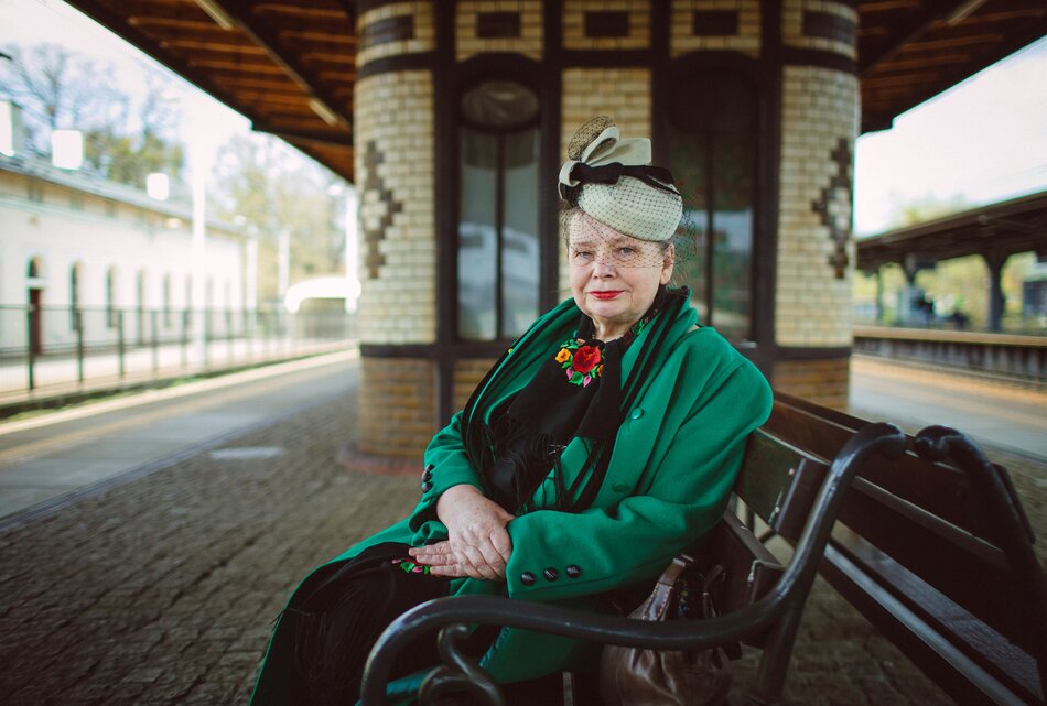 Na zdjęciu kobieta w dojrzałym wieku, ubrana w zieloną marynarkę, na głowie gustowna wolaka. Kobieta siedzi na ławce na starym dworcu kolejowym.