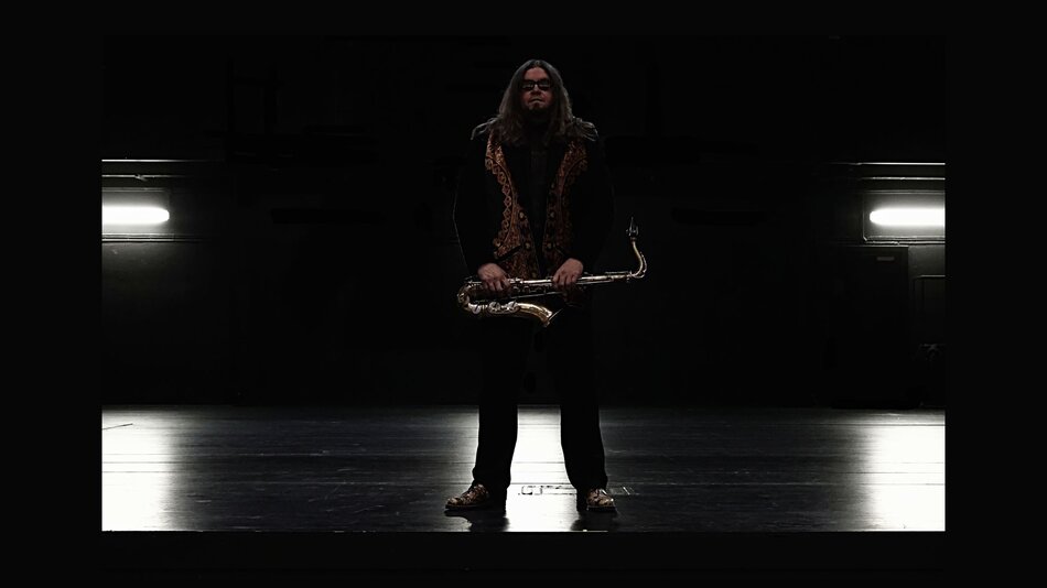 Na zdjęciu mężczyzna o długich włosach, w ciemnych okularach, stojący na scenie, trzymający saksofon.