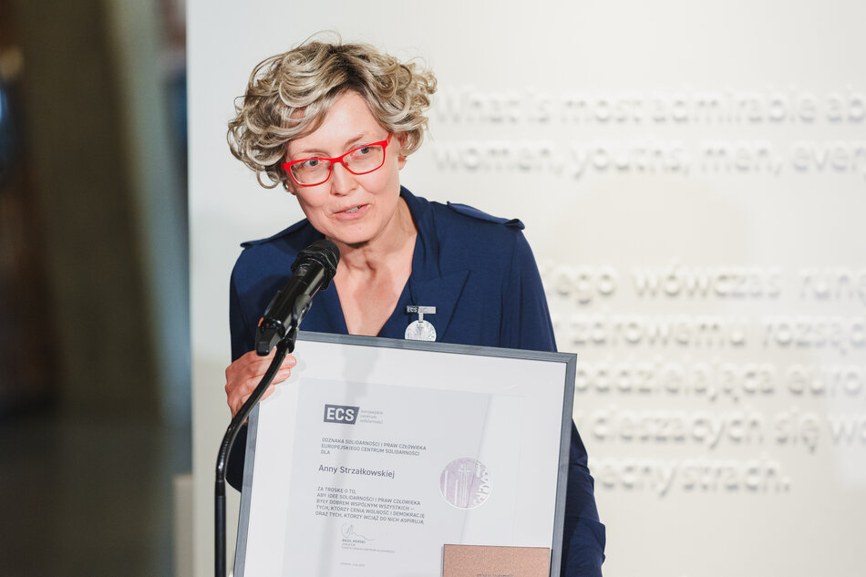 Na zdjęciu widzimy Annę Strzałkowską trzymającą w dłoniach oprawione w ramę odznaczenie przyznane przez Europejskie Centrum Solidarności. Ubrana w granatową sukienkę i czerwone okulary, przemawia do mikrofonu podczas uroczystości wręczenia Odznaki Solidarności i Praw Człowieka.