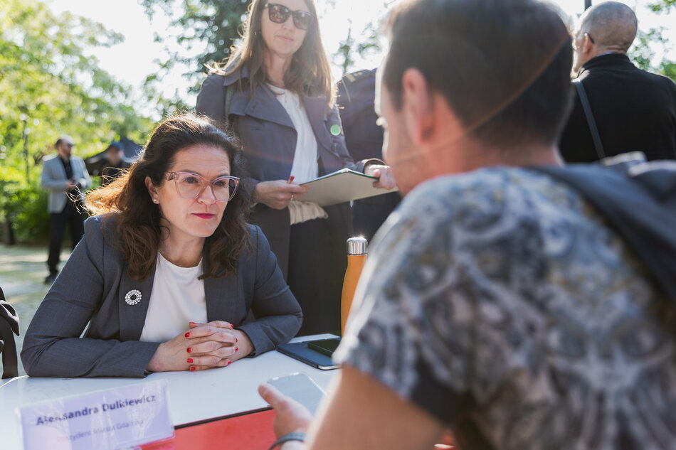 na zdjęciu kobieta w średnim wieku, w szarej marynarce, siedzi przy stole, patrzy na mężczyznę który siedzi naprzeciw niej