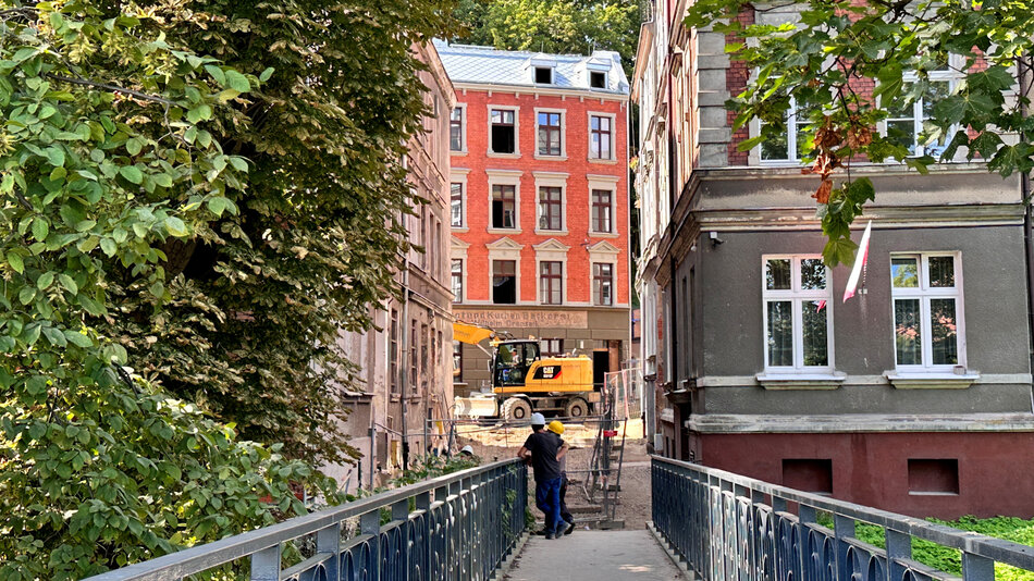 Zdjęcie przedstawia alejkę między kamienicami, prowadzącą do remontowanego budynku z cegły, przed którym stoi koparka i dwóch robotników w kaskach. Po bokach widać bujną zieleń oraz starsze kamienice, nadające miejscu klimatyczny charakter. 