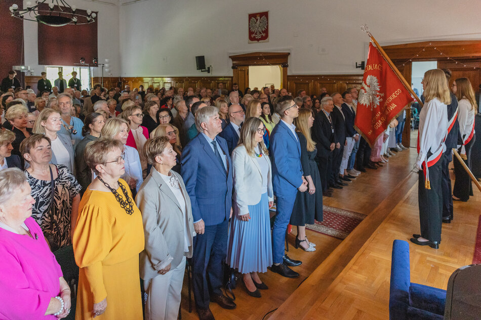 Uczestnicy uroczystości stoją w sali z pocztem sztandarowym, w centrum widoczna jest kobieta w białym żakiecie. Sala jest wypełniona gośćmi, co sugeruje ważne wydarzenie, jakim jest jubileusz