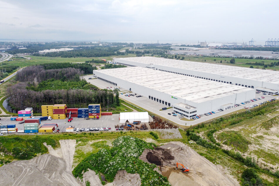 Na zdjęciu widać duży kompleks magazynowy zlokalizowany w pobliżu portu morskiego, otoczony terenami zielonymi i infrastrukturą logistyczną. Przed magazynem znajdują się kontenery, place manewrowe, parkingi oraz miejsce budowy z pracującą koparką, co wskazuje na rozwijającą się działalność przemysłową i transportową. 