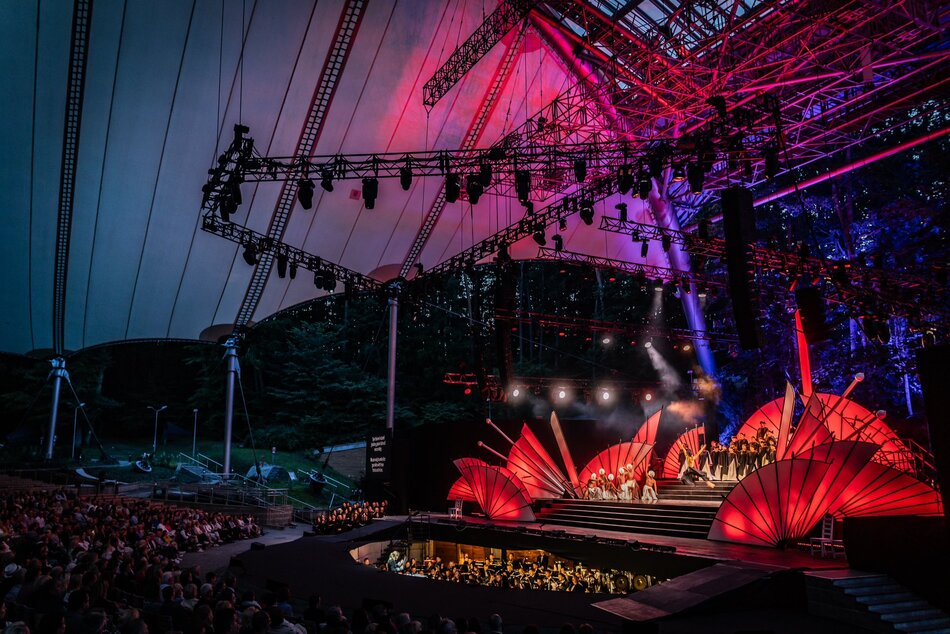 Plenerowa scena operowa znajduje się pod ogromnym zadaszeniem, z imponującą scenografią w kształcie czerwonych wachlarzy i intensywnym oświetleniem. Widownia oraz orkiestra znajdują się na niższym poziomie, a spektakl rozgrywa się na tle drzew, nadając wydarzeniu wyjątkową atmosferę
