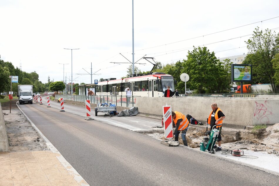 Na zdjęciu widać trwające prace drogowe przy torowisku tramwajowym — dwóch robotników w pomarańczowych kamizelkach wykonuje roboty przy krawędzi jezdni, otoczonej barierkami ostrzegawczymi. W tle zatrzymał się tramwaj, z którego wysiadają pasażerowie, a obok widoczna jest ciężarówka i znaki informujące o robotach drogowych. 