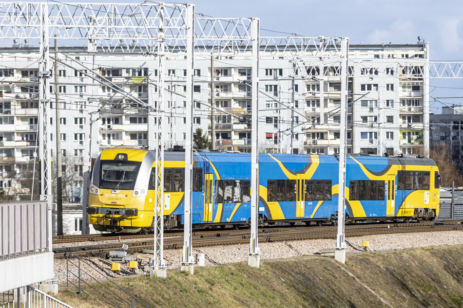 Na zdjęciu widoczny jest pociąg Pomorskiej Kolei Metropolitalnej (PKM) w charakterystycznych barwach żółto-niebieskich, poruszający się po estakadzie wśród infrastruktury trakcyjnej. W tle znajdują się bloki mieszkalne, co sugeruje, że skład przejeżdża przez miejską zabudowę Gdańska. 