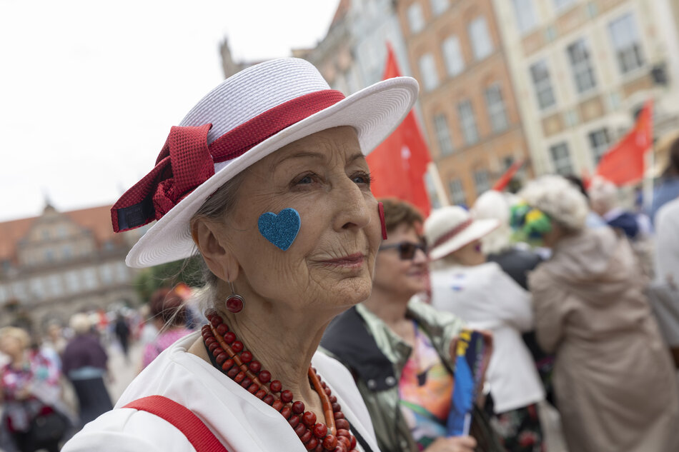 Na zdjęciu starsza kobieta w eleganckim białym kapeluszu z czerwonymi akcentami i niebieskim serduszkiem na policzku uczestniczy w kolorowej paradzie. W tle widać tłum innych uczestników, a atmosfera wydarzenia jest radosna i pełna dumy. 