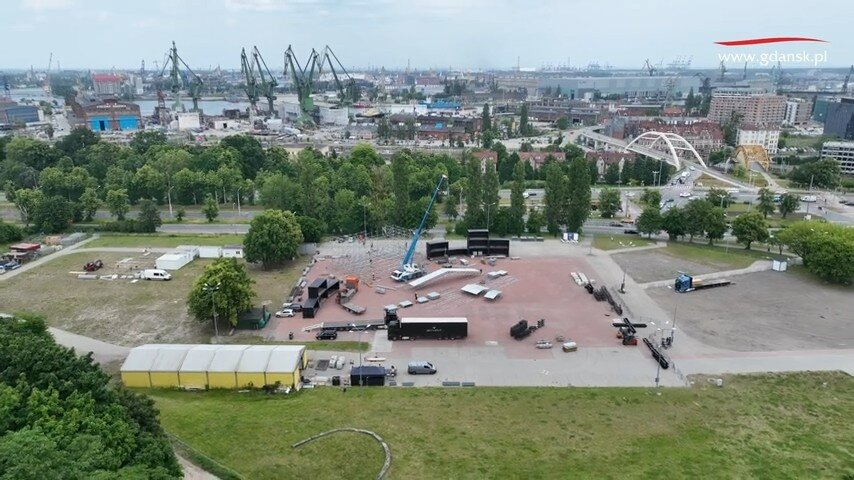 Na zdjęciu widać przygotowania do dużego wydarzenia plenerowego w Gdańsku — montowany jest duży podest sceniczny oraz infrastruktura techniczna. W tle rozciąga się panorama Stoczni Gdańskiej z charakterystycznymi dźwigami oraz miejską zabudową. 