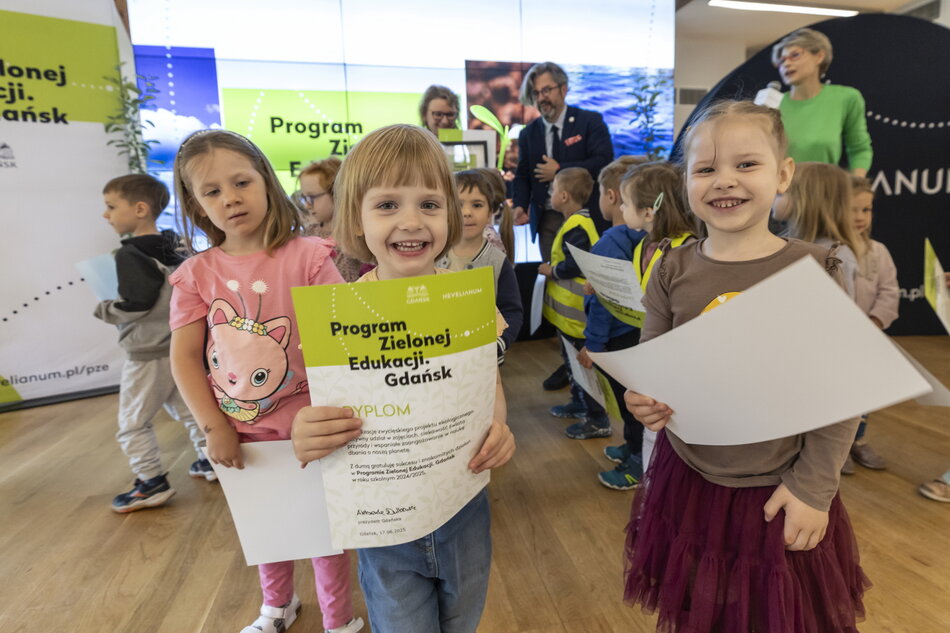 Na zdjęciu dzieci z Przedszkola nr 42 radośnie prezentują dyplomy otrzymane podczas finału Programu Zielonej Edukacji w Gdańsku. W tle widać organizatorów wydarzenia oraz dekoracje związane z ekologicznym charakterem programu. 