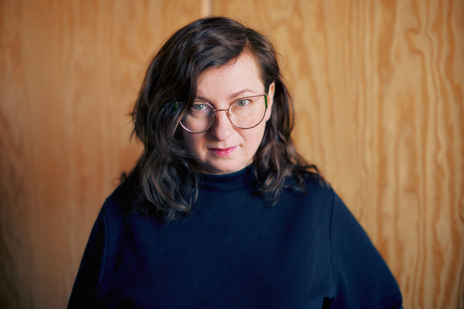 Na zdjęciu kobieta z ciemnymi włosami za ramiona, patrząca na wprost, w tle ściana.