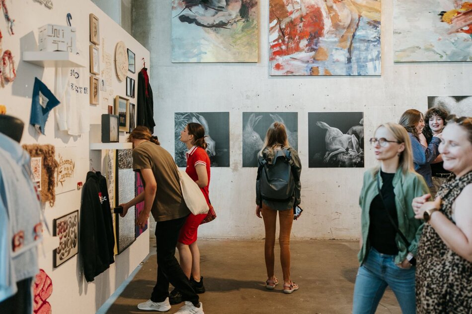 W tej części wystawy widać różnorodne prace: od haftów, przez grafikę, po rysunki zwierząt na ścianie. Kilka osób dokładnie przygląda się detalom ekspozycji, tworząc żywy klimat zainteresowania