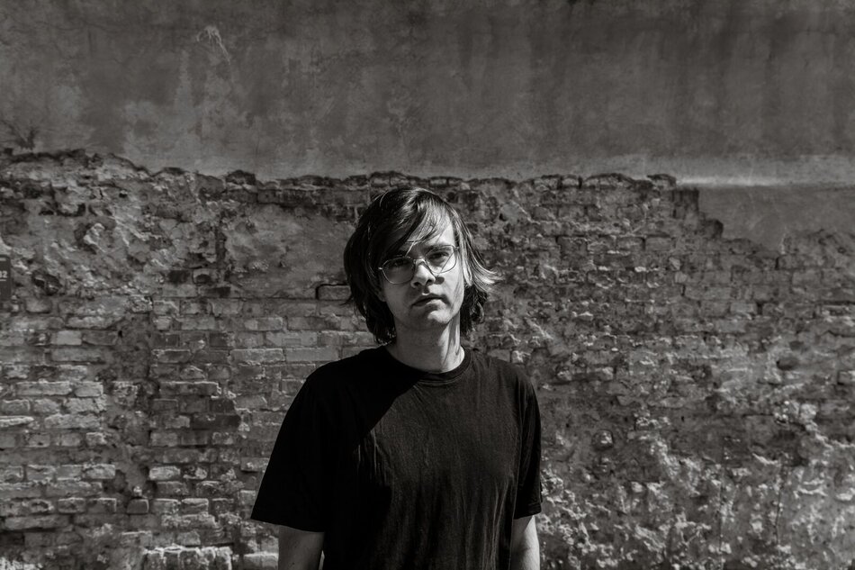 Na zdjęciu mężczyzna z włosami za uszy, ubrany w czarny T-shirt, na tle zniszczonej ściany.