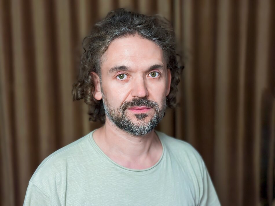 Na zdjęciu mężczyzna z kręconymi włosami i zarostem. Na tle brązowej kotary.