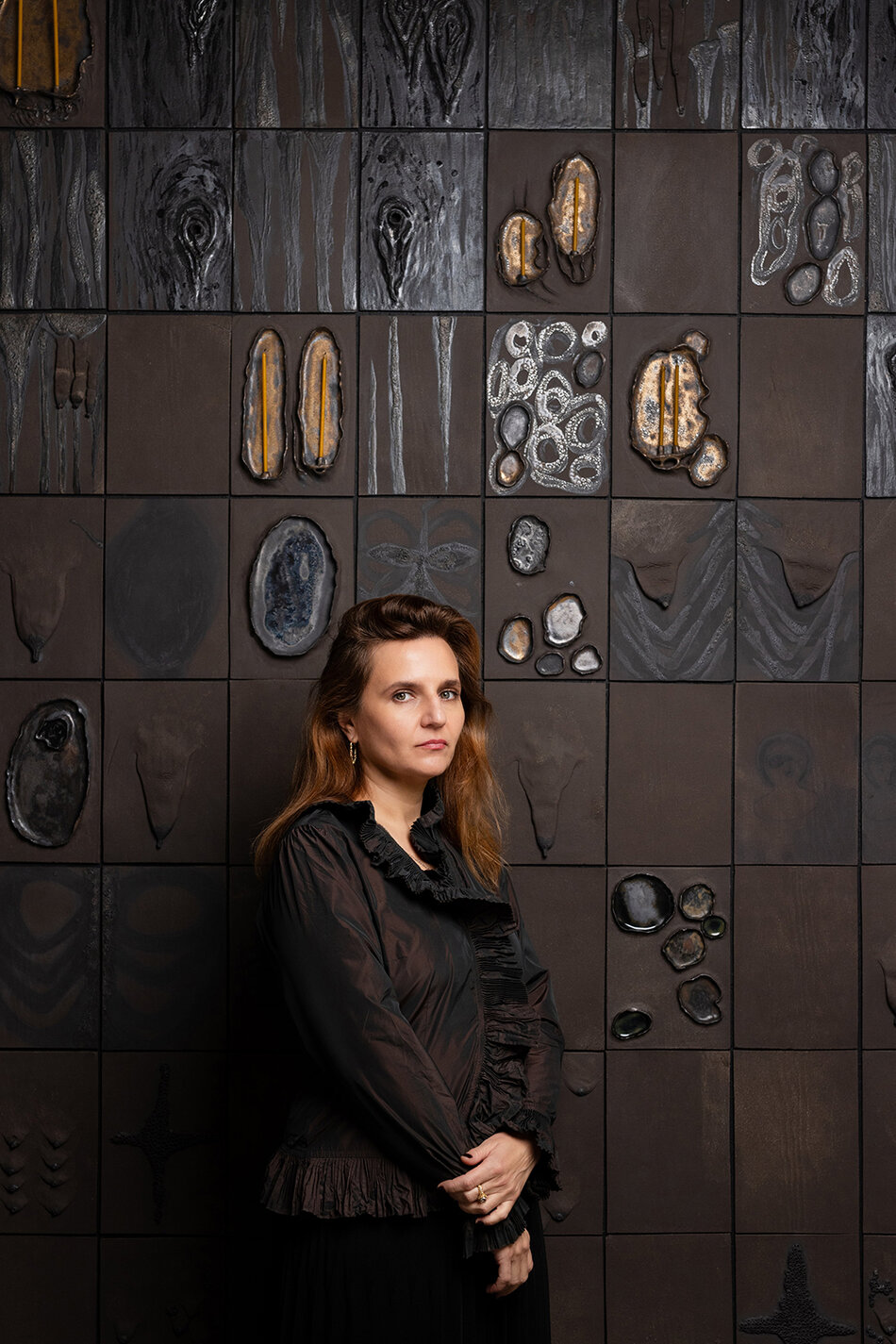Na zdjęciu kobieta z włosami za ramiona, w ciemnym stroju, na tle artystycznej ściany z wieloma nieregularnymi kształtami.