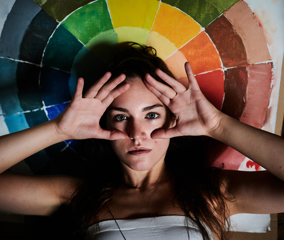 Na zdjęciu kobieta z przyłożonymi do twarzy rękoma, na tle wielobarwnego obrazu.