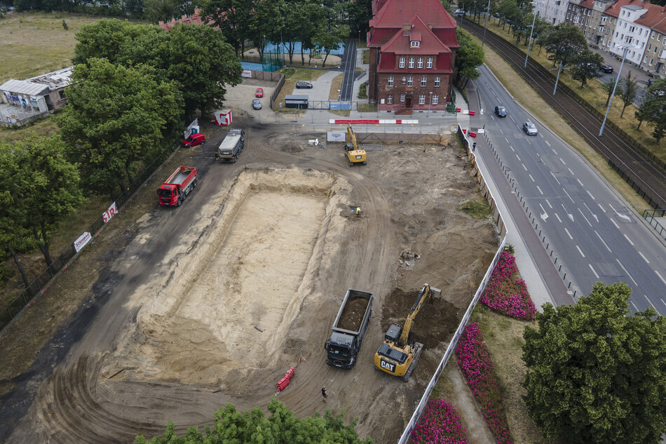 zdjęcie z drona, widać plac budowy a na nim dwie żółte koparki i kilka ciężarówek, jest też wykopany dół w ziemi