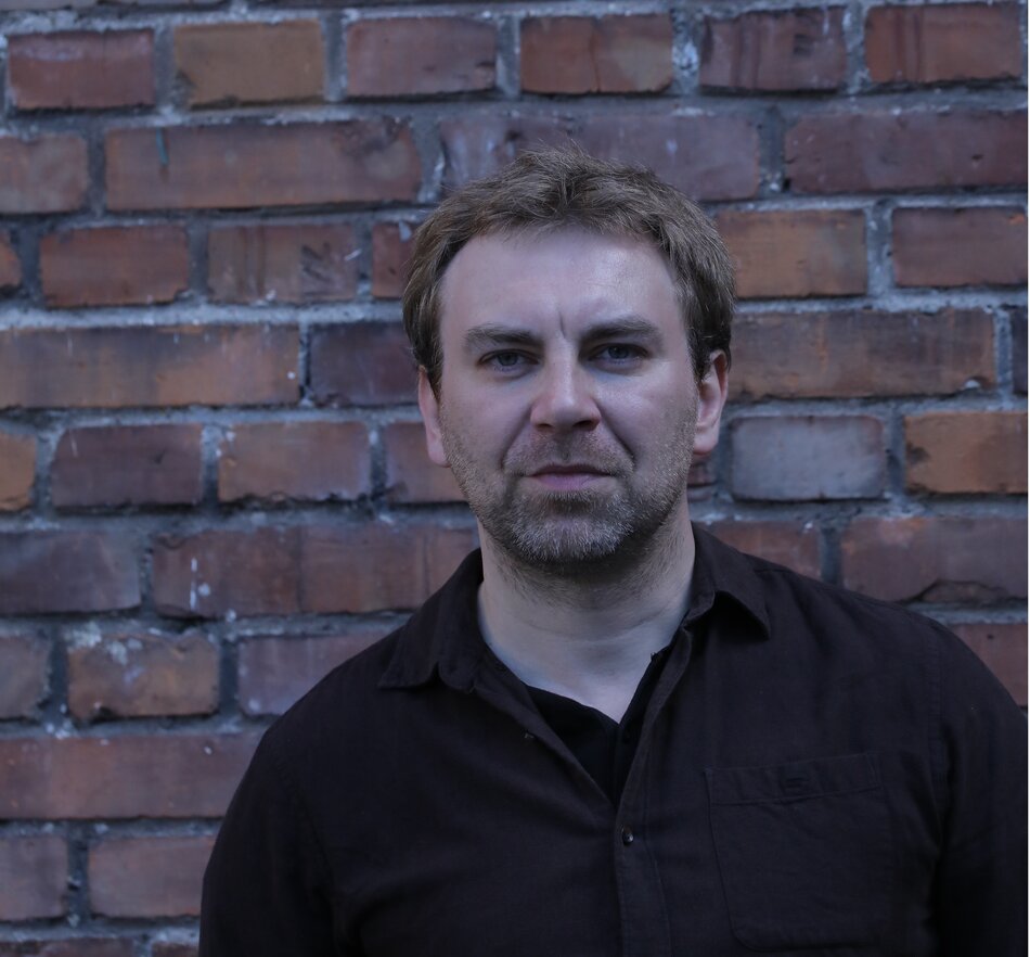 Na zdjęciu mężczyzna z krótkimi włosami i lekkim zarostem na tle ceglanej ściany.