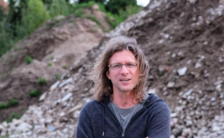 Na zdjęciu mężczyzna z włosami za ramiona, w okularach, na tle nasypów z kamieni i ziemi.