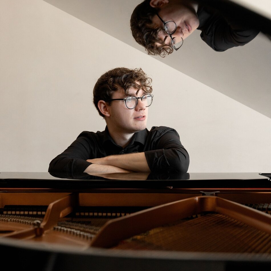 Na zdjęciu mężczyzna w okularach oparty o otwartą klapę fortepianu.