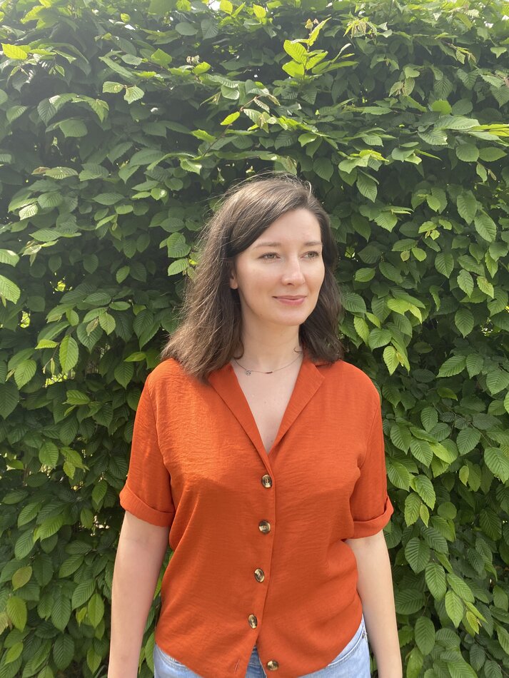 Na zdjęciu kobieta z włosami za ramiona, ubrana w pomarańczową letnią bluzkę, na tle zielonych liści.